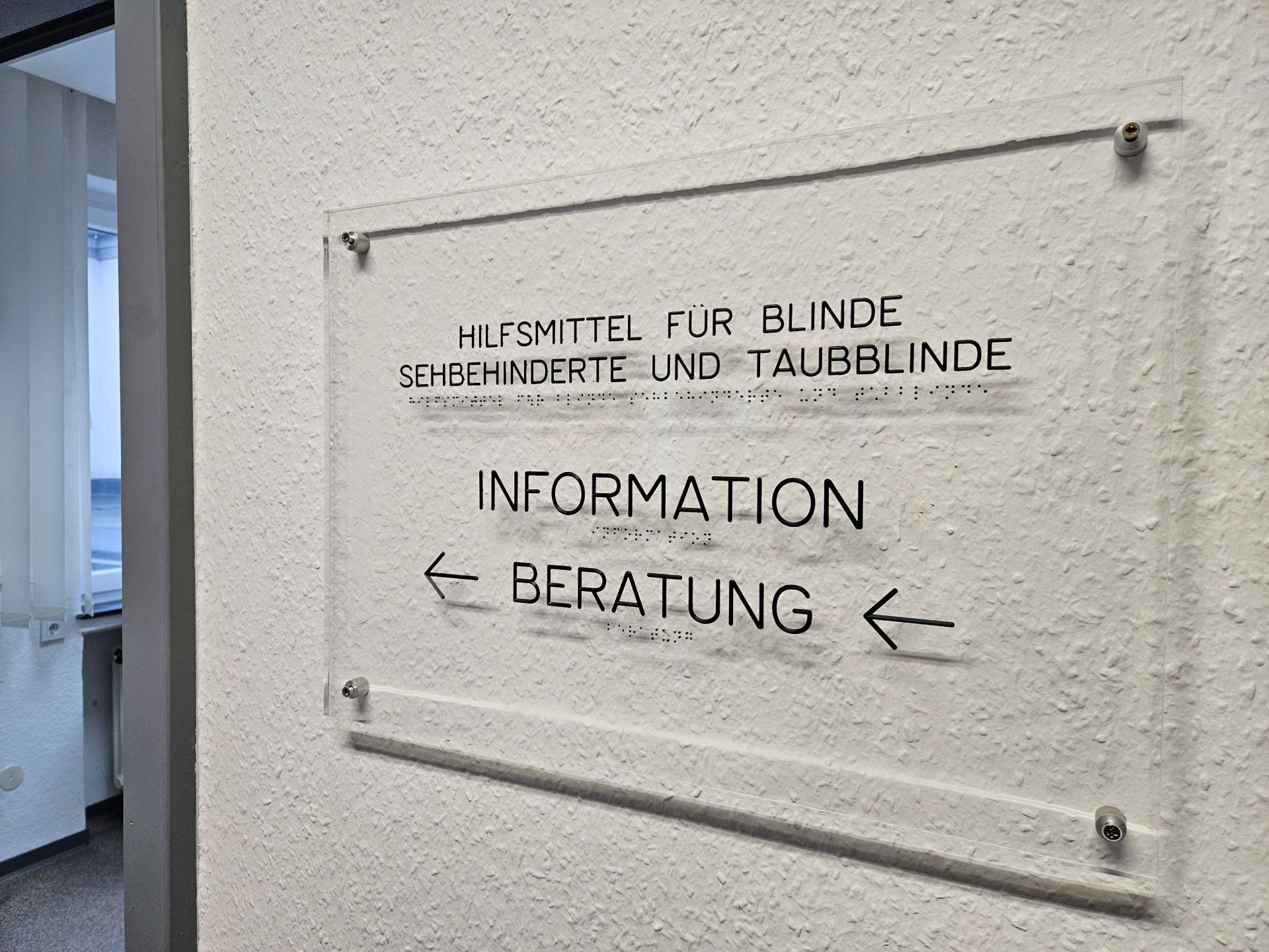 Ein durchsichtiges Hinweisschild an einer weißen Wand mit Raufasertapete. Auf dem Schild steht in Schwarzschrift und Braille Information, Beratung.