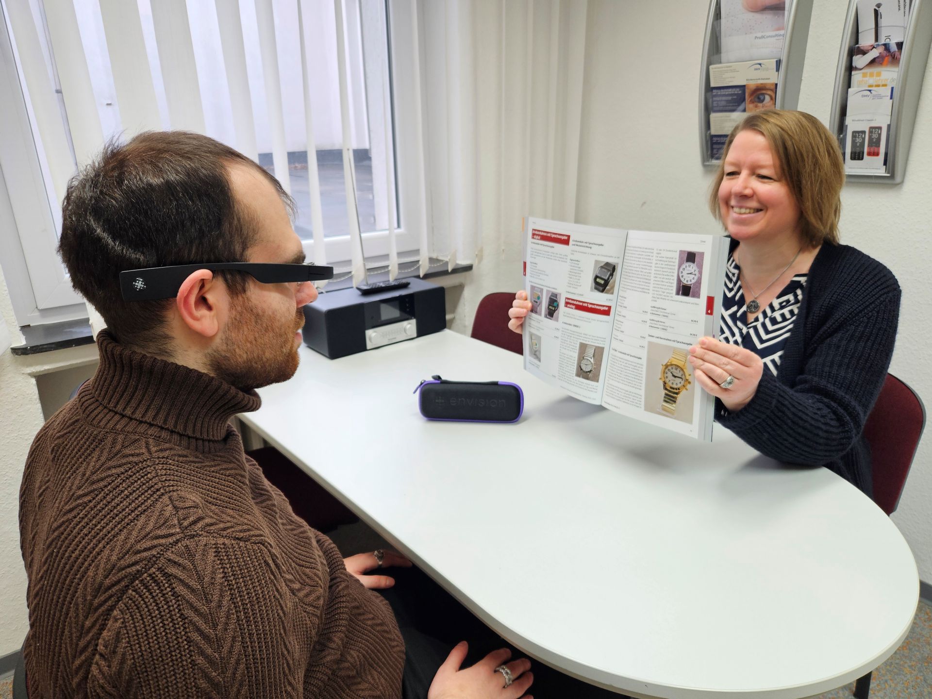 Verkaufsgespräch an einem weißen Tisch. Eine freundlich lächelnde Frau zeigt einen Katalog hoch. Der junge Mann ihr gegenüber trägt eine spezielle Brille und blickt auf den Katalog.
